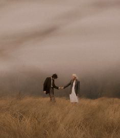 a man and woman are holding hands in a field with tall grass under a cloudy sky