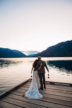 a bride and groom standing on a dock in front of the water at sunset with their arms around each other