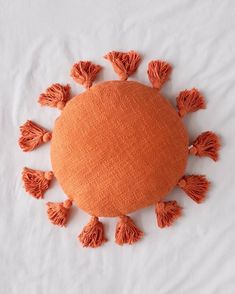 an orange round cushion with tassels on the top and bottom, sitting on a white surface