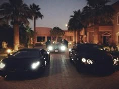 two black sports cars parked in front of a house at night with palm trees on both sides