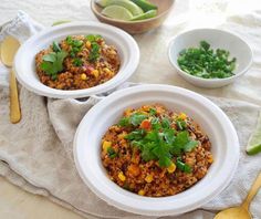 two bowls filled with rice and vegetables on top of a white cloth next to silverware