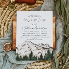 a wedding card with mountains and trees on it, surrounded by greenery in the background