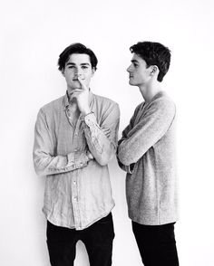 two men standing next to each other in front of a white wall with their hands on their chins