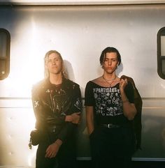 two young men standing next to each other in front of a silver train with their hands on their hips