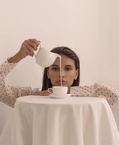 a woman sitting at a table drinking from a white coffee cup and pouring water into her teacup