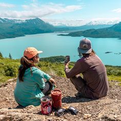 two people sitting on the side of a mountain taking pictures with their cell phones and camera