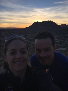 two people taking a selfie in front of a mountain range at sunset with the sun setting behind them