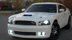 a white car parked in a parking lot next to some trees and bushes with lights on