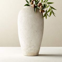 a white vase with some green leaves and flowers in it on a countertop next to a wall