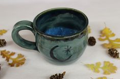 a green mug with a crescent and stars on it sitting next to some pine cones