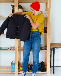 a woman in yellow shirt and blue jeans holding up a black jacket on wooden shelves