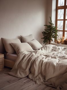 an unmade bed in front of a window with candles and a christmas tree next to it