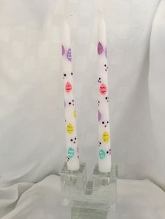 two white candles with colorful designs on them sitting in small glass holders, one is lit and the other is turned upside down