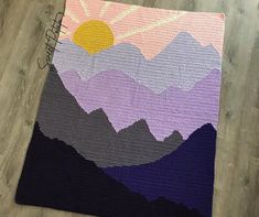 a crocheted blanket with mountains and sun in the background on a wooden floor