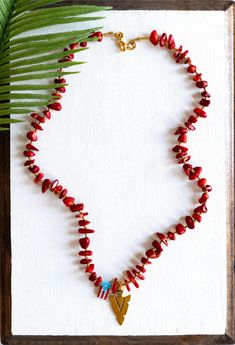 a red beaded necklace with a gold heart charm hangs from a wooden frame next to a palm leaf