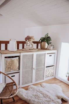 a room with white furniture and plants on top of the cabinet, in front of a window