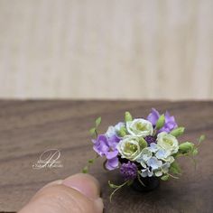 a hand is holding a small vase with purple and white flowers in it on a wooden table