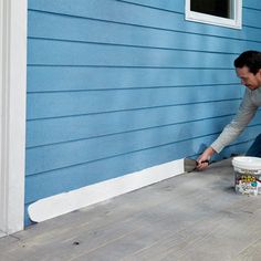 a man painting the side of a house with blue paint and a white bucket next to it