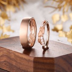 two wedding rings with leaves on them sitting on top of a wooden stand in front of gold foil