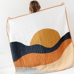 a woman is holding up a quilt that looks like mountains and the sun in the sky