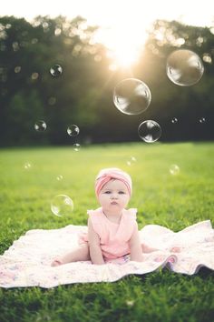 a baby sitting on a blanket in the grass with soap bubbles floating above her head