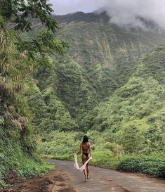 a woman walking down a road with a surfboard in her hand and mountains in the background