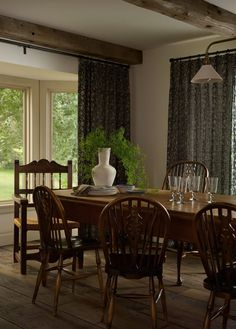 a dining room table with chairs and a vase on top of it in front of two windows