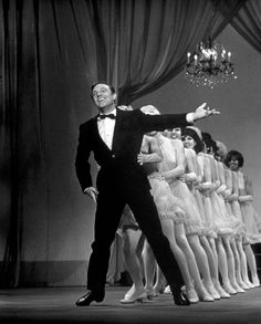 a man in a tuxedo standing next to a group of women on stage