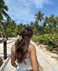 a woman walking down a walkway with palm trees in the background and a white flower in her hair