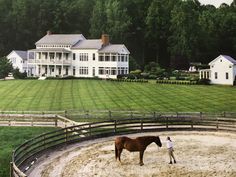 a man standing next to a brown horse in front of a large white house on top of a lush green field