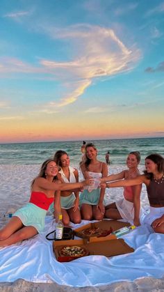 four women are sitting on the beach and toasting with each other as the sun sets