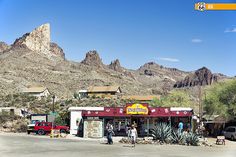 people are standing in front of a small store on the side of a road with mountains in the background