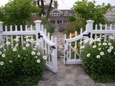 an open white gate leading into a garden with flowers and trees in front of the house