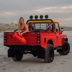 a woman sitting in the back of a red truck on top of a sandy beach