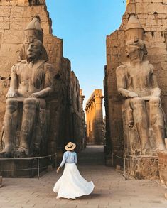 a woman in a white dress and straw hat walking through an ancient city with statues