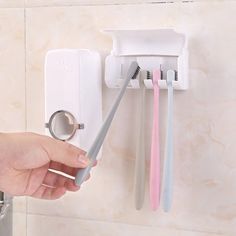 a person is holding two toothbrushes in front of a wall mounted toothpaste dispenser