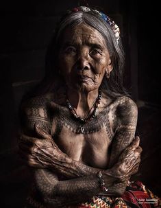 an old woman with tattoos on her body and chest is sitting in the dark, looking at the camera