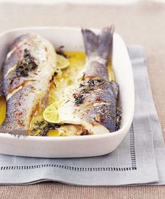 two fish in a white dish with lemon slices and herbs on the side, sitting on a napkin