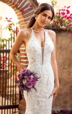 a woman in a wedding dress standing next to a brick wall and holding a purple bouquet