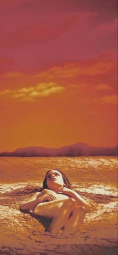 a woman laying on top of a sandy beach under a red and orange sky at sunset