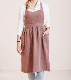 a woman standing in front of a white wall wearing a brown apron and blue jeans