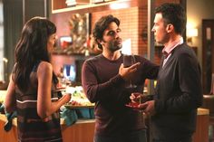 two men and a woman standing in a kitchen talking to each other while holding wine glasses