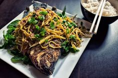 a white plate topped with fish covered in noodles and veggies next to a bowl of rice