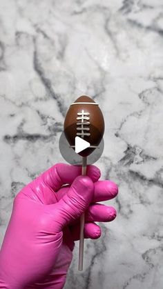 a person in pink gloves holding up a chocolate football on a toothpick with a stick sticking out of it