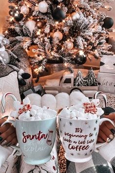 two coffee mugs with marshmallows in front of a decorated christmas tree