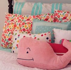 a pink whale pillow on top of a bed next to pillows with flowers and stripes