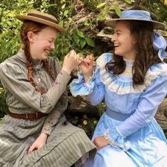 two women in dresses and hats are sitting on a bench eating fruit together, with one woman smiling at the camera