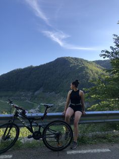 a woman sitting on the side of a road next to a bike with mountains in the background