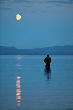 a man standing in the water with a fishing pole at night, while the moon is setting