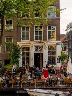 people are sitting at tables on the water in front of a building with many windows